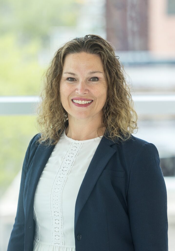 A woman with curly brown hair, wearing a navy blazer over a white blouse, smiles at the camera. The background is blurred with hints of greenery and buildings.