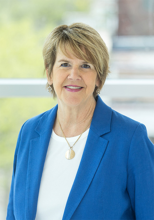 A woman with short, light brown hair smiles at the camera. She is wearing a blue blazer over a white top, a gold necklace, and earrings. The background is softly blurred with greenery and a window.