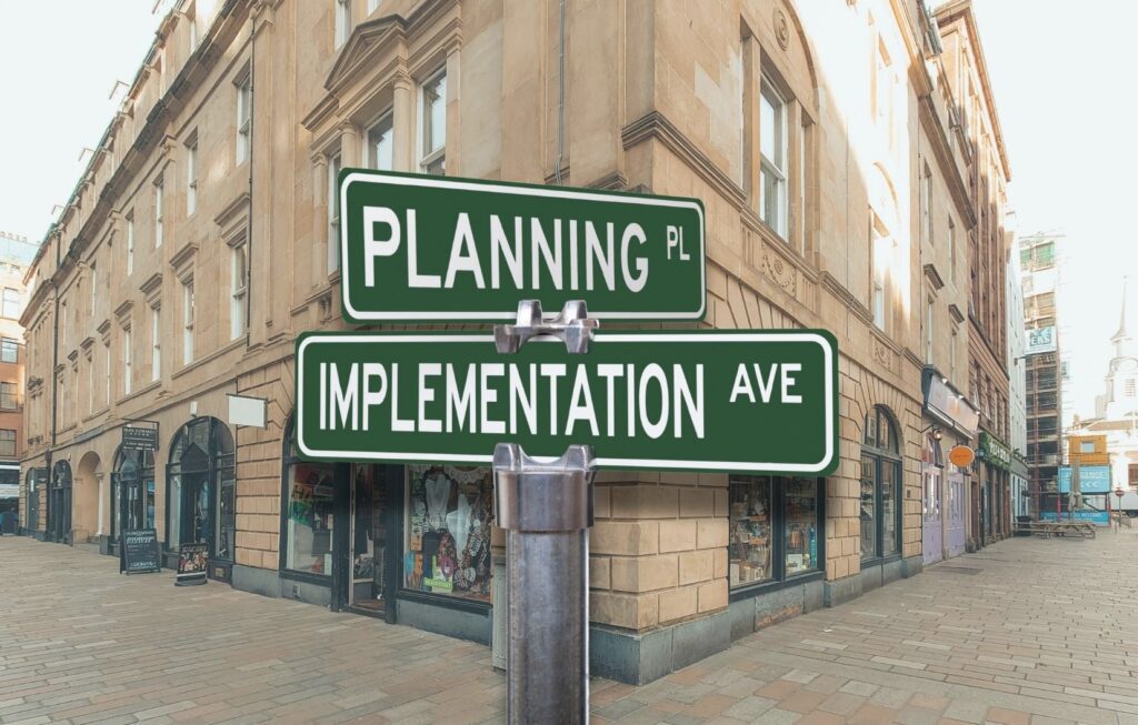 Two green street signs at a corner read PLANNING PL and IMPLEMENTATION AVE, attached to a pole in front of a beige building on a quiet city street.