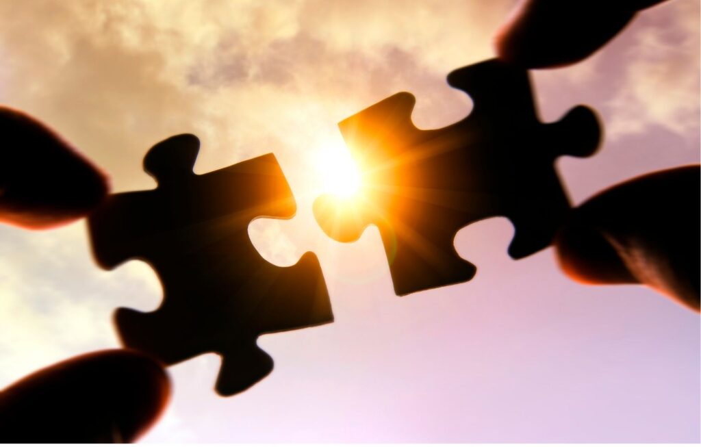 Two hands holding puzzle pieces up to the sky, with sunlight shining between the pieces, suggesting connection or completion.